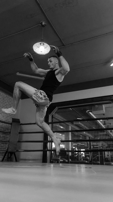Black and white photo of an athlete in midair performing a jumping kick in a boxing ring.