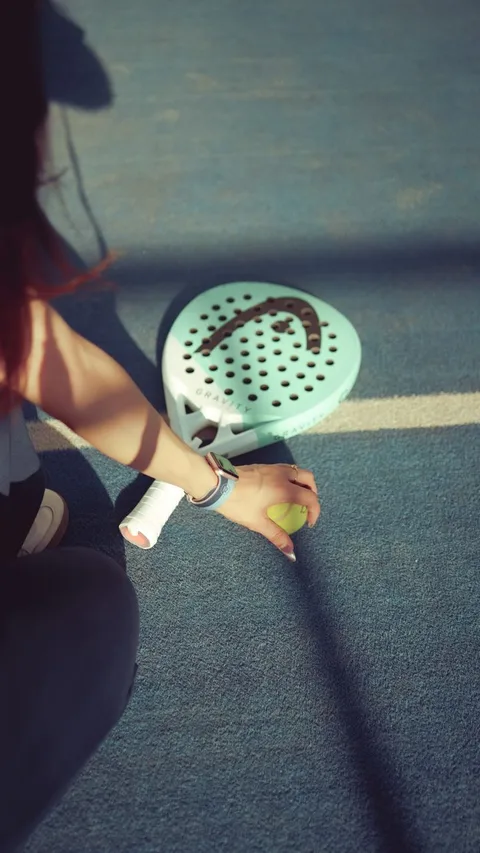 A young adult prepares for a padel match, holding a racket and ball on a sunlit court.