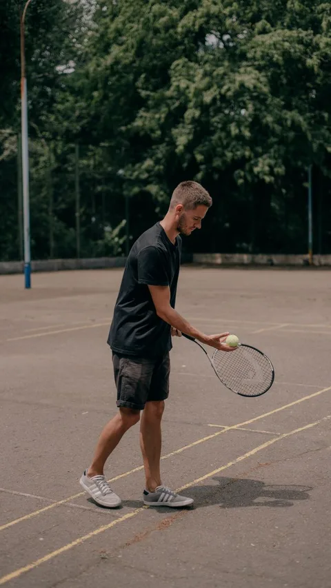 A young man in a black shirt playing tennis on an outdoor court surrounded by trees.