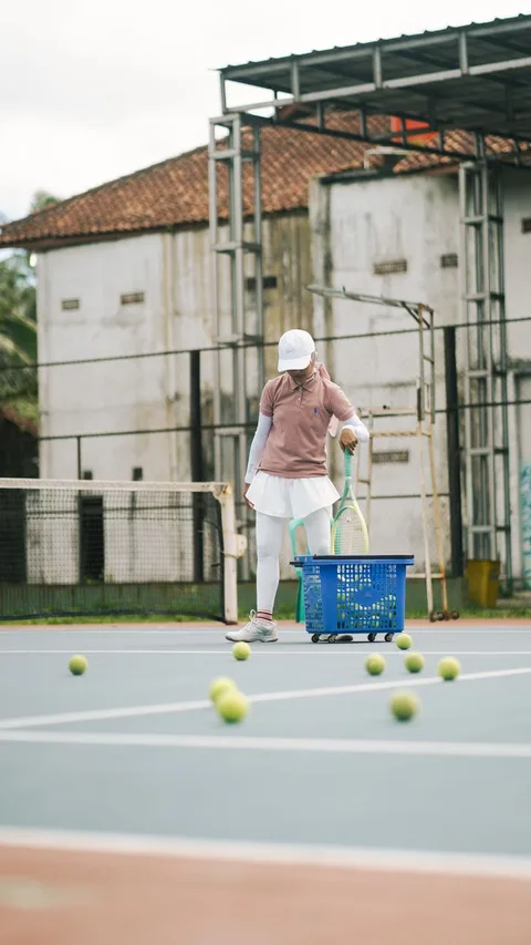 A tennis player collects balls on an outdoor court, focusing on training with scattered tennis balls.